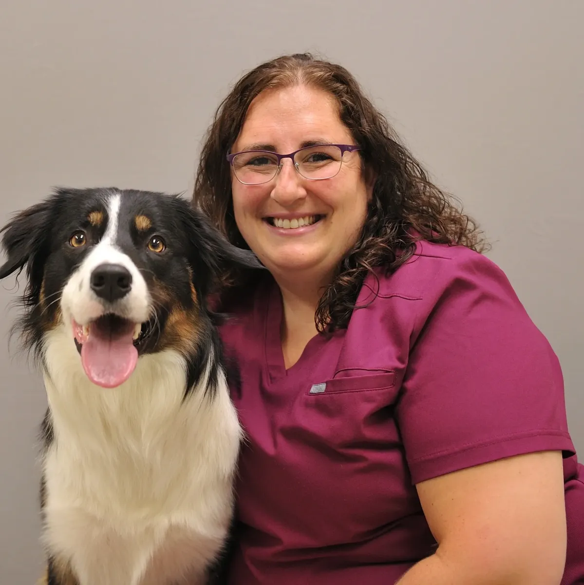 Woman in maroon scrubs and glasses smiling with a black, white, and tan dog with its tongue out against a gray background