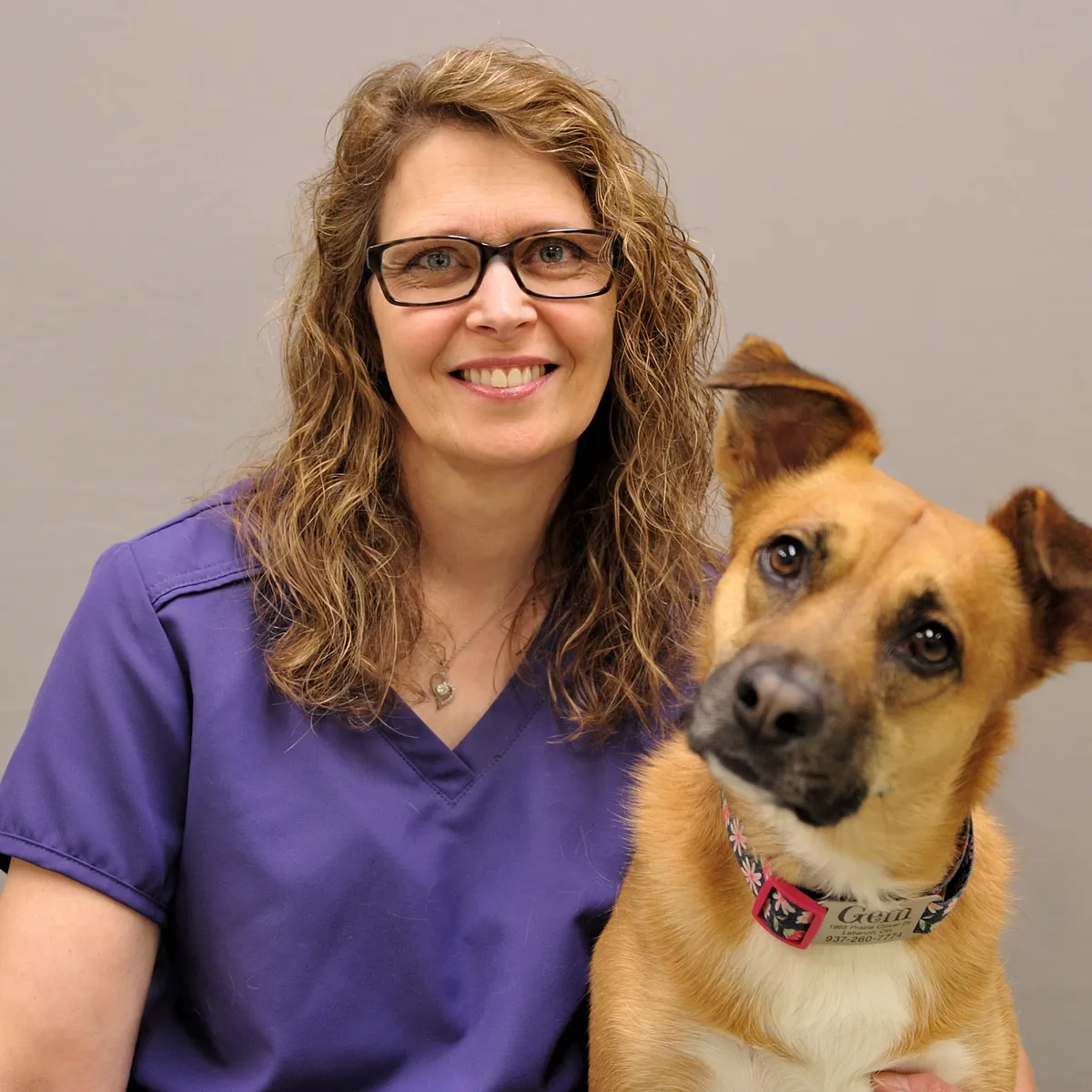 Woman in purple scrubs wearing glasses smiles while holding a tan and white dog wearing a red collar, representing compassionate pet care at a Centerville Ohio veterinary practice