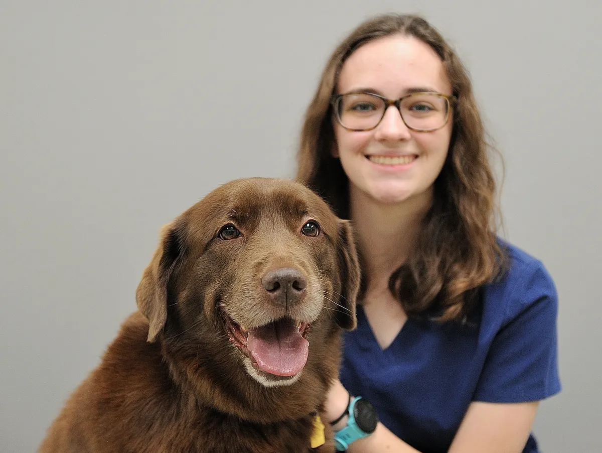 Young woman wearing glasses and blue scrubs smiling while posing with a brown Labrador Retriever dog wearing a teal collar