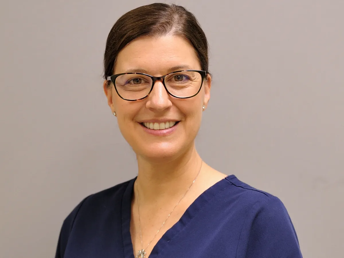 Woman with dark hair and glasses wearing a navy blue top, smiling at the camera against a gray background