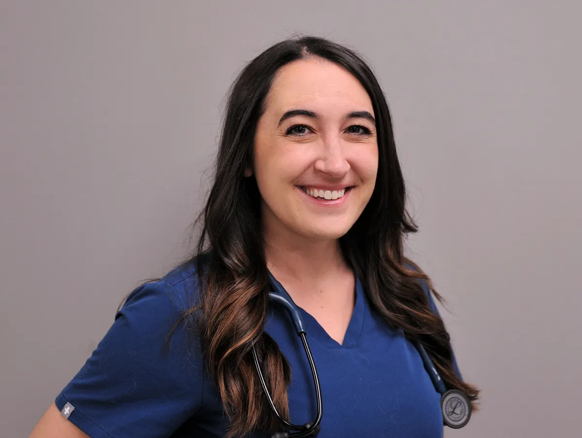 Healthcare professional in blue scrubs with stethoscope smiling at camera against gray background