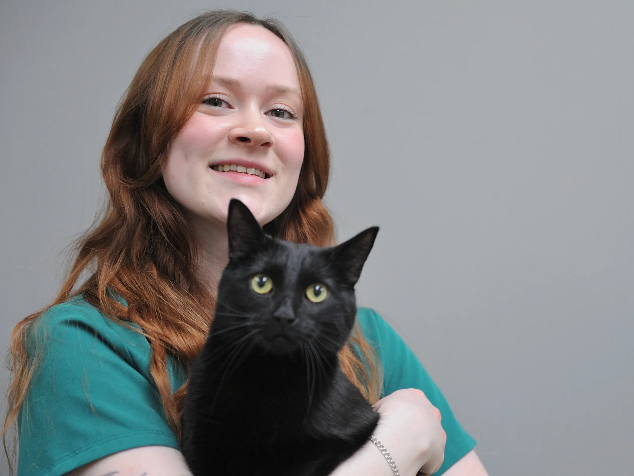 Young woman with reddish-brown hair wearing a teal shirt, smiling while holding a black cat with yellow eyes