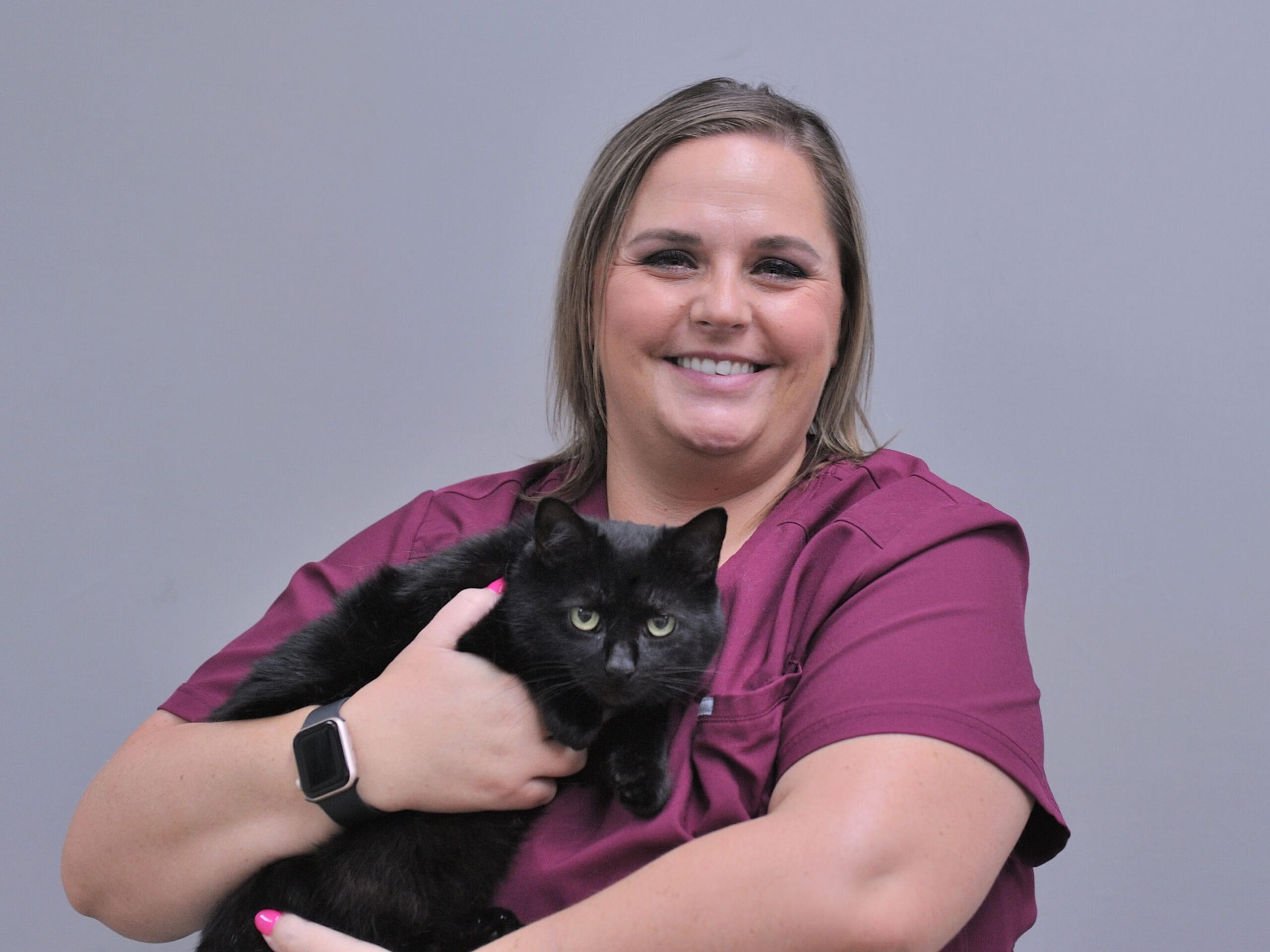 Smiling veterinarian in maroon scrubs holding a black cat with yellow eyes against a gray background