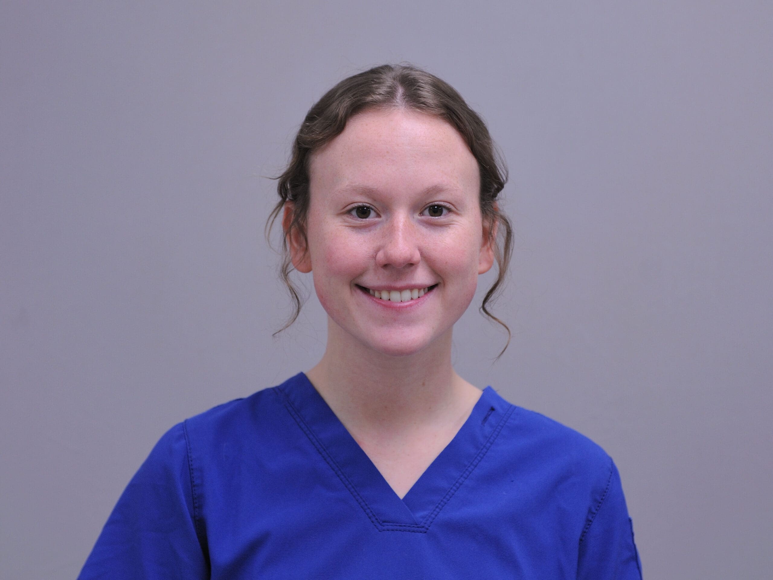Smiling woman with brown hair wearing blue medical scrubs against gray background