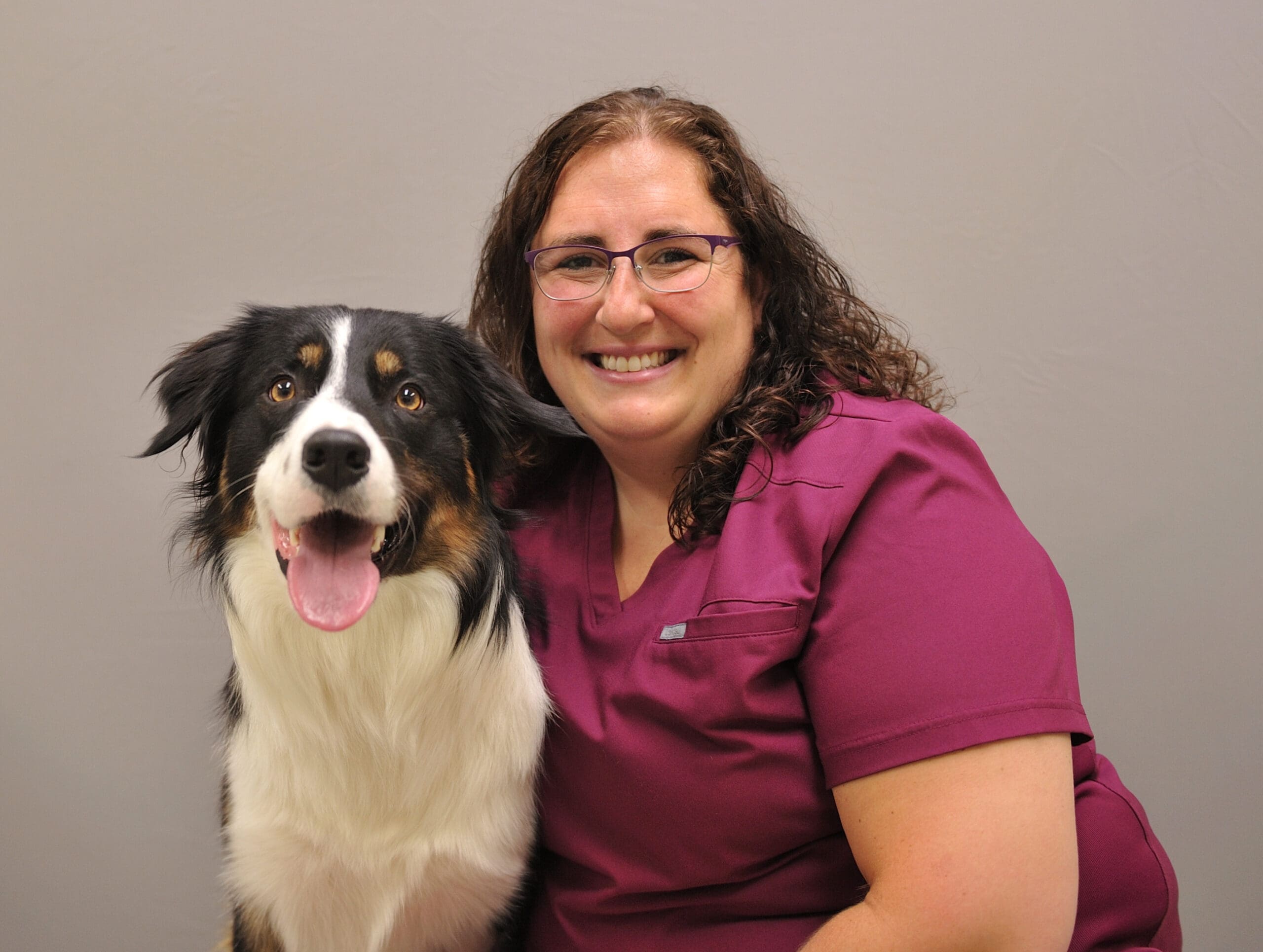 Smiling veterinarian in maroon scrubs with glasses posing beside a black and white dog with tongue out against a gray background