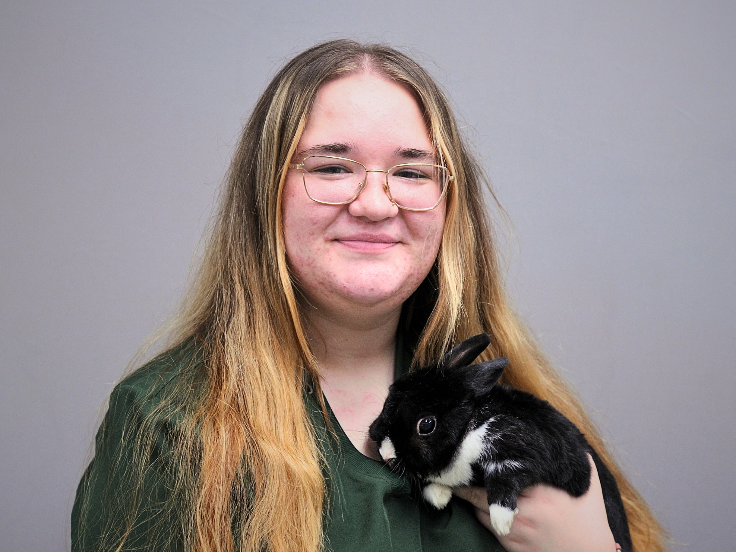 Young woman with long reddish-brown hair and glasses wearing a dark green shirt, holding a black and white rabbit against her chest